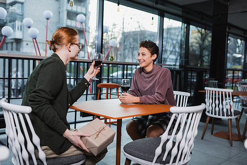 Young man holding present near smiling girlfriend with wine in hotel cafe