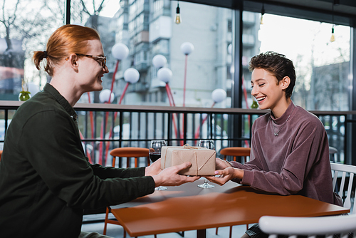 Smiling man giving present to girlfriend near wine in hotel cafe