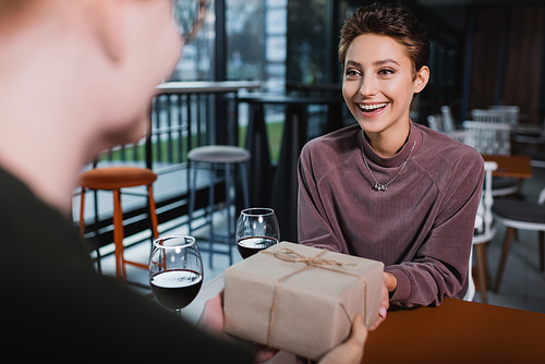 Excited woman holding present near wine and blurred boyfriend in hotel cafe