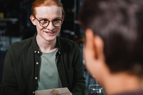 Smiling man in eyeglasses holding present near wine and blurred girlfriend in hotel