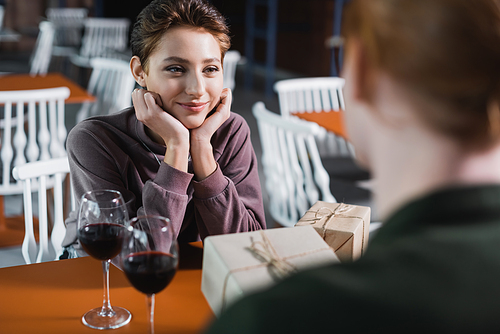 Young woman looking at blurred boyfriend with gift near wine in hotel cafe