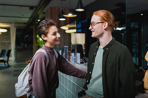 Smiling man talking to girlfriend with backpack near reception in hotel