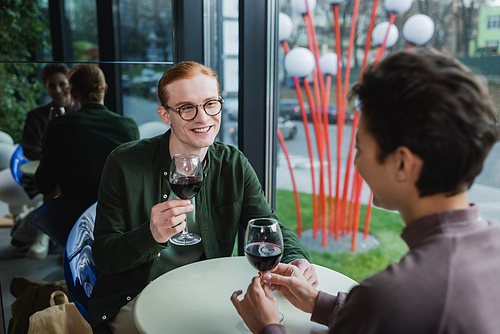 Smiling red haired man holding glass of wine near blurred girlfriend in hotel