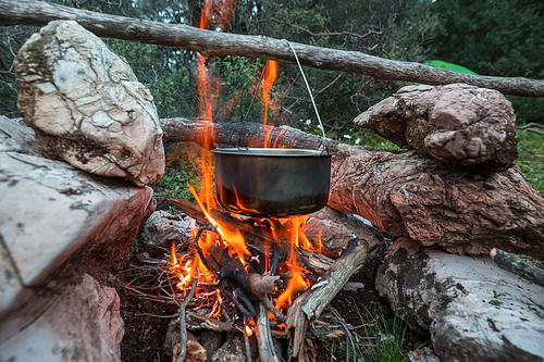 Touristic kettle on fire of burning campfire in camping in the hike. Cooking food in forest on wooden firewood.