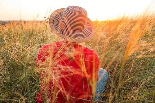 a girl in a hat sitting in the grass in a meadow at sunset