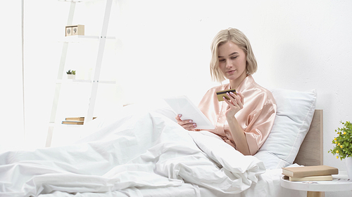 blonde woman holding digital tablet and credit card while doing online shopping in bed