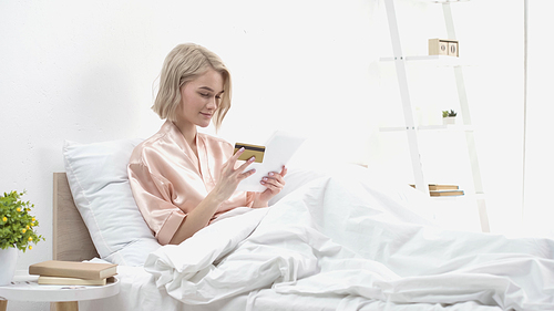 smiling woman holding digital tablet and credit card while doing online shopping in bed