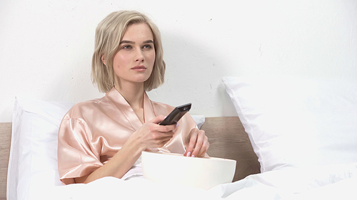 blonde woman sitting with bowl while holding remote controller and watching movie