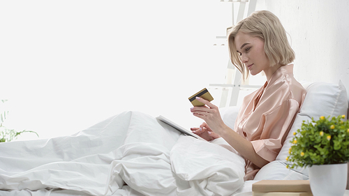 side view of blonde woman holding digital tablet and credit card in bed