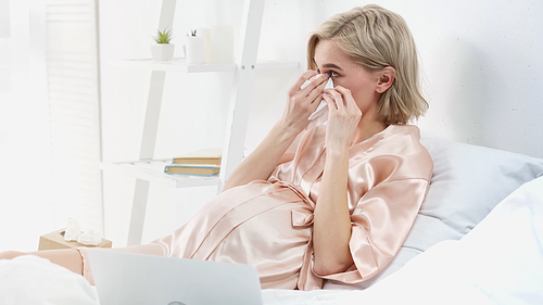 sad blonde pregnant woman wiping tears with napkin while watching movie on laptop in bedroom