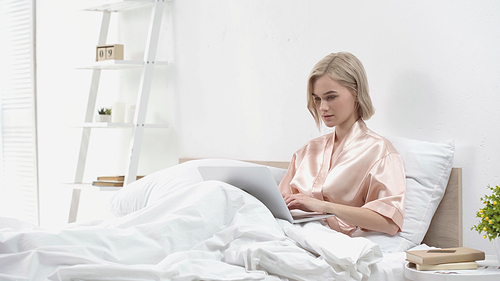 young blonde freelancer using laptop while sitting in bed