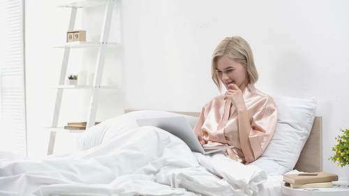 smiling blonde freelancer using laptop while sitting in bed