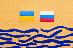 Top view of Ukrainian and russian flags above paper sea on textured yellow background
