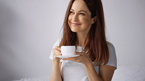 joyful woman holding coffee cup while looking away on grey background