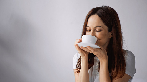 pleased woman with closed eyes enjoying morning coffee on grey background