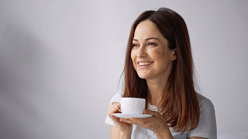 cheerful woman looking away while holding white coffee cup on grey background