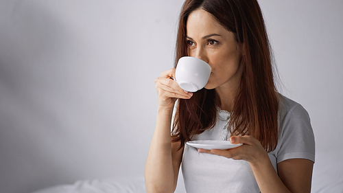 brunette woman drinking coffee from white cup in morning on grey background