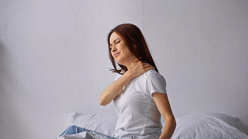 brunette woman with closed eyes sitting on bed and touching painful neck