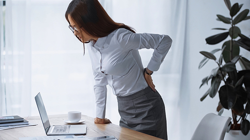 brunette woman touching painful back while standing at work desk