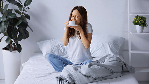 pleased woman in pajamas sitting on bed and smelling aromatic coffee