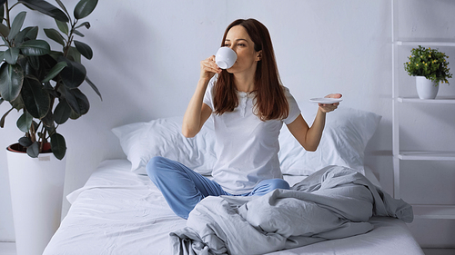 brunette woman in pajamas drinking morning coffee while sitting on bed
