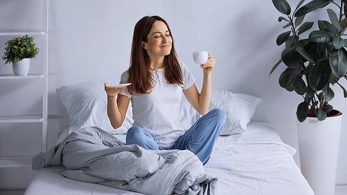 smiling woman in pajamas holding coffee cup while sitting on bed with closed eyes