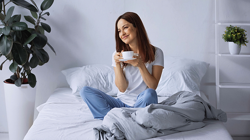 happy brunette woman in pajamas looking away while sitting on bed with cup of coffee