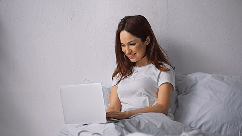 brunette woman smiling while using laptop on bed at home