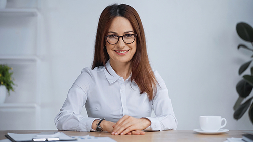 brunette businesswoman in eyeglasses sitting at workplace and smiling at camera