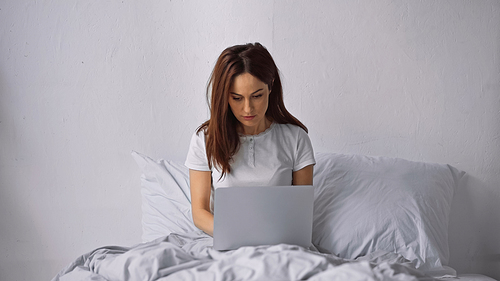 brunette woman sitting on bed at home and working on laptop