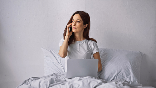 brunette woman sitting on bed with laptop and talking on mobile phone
