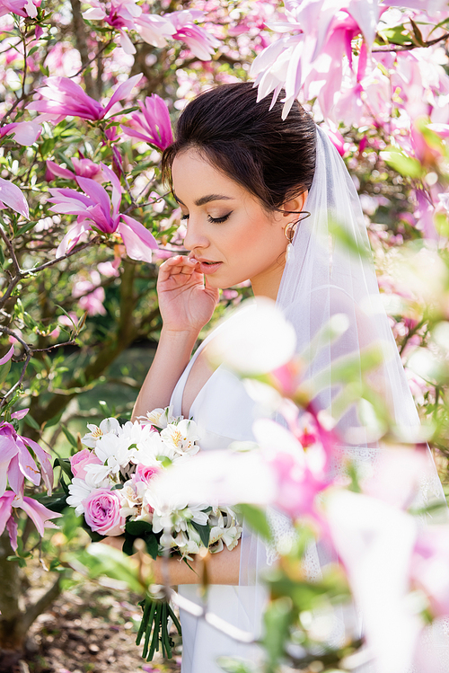 Young bride in veil standing near blooming magnolia tree