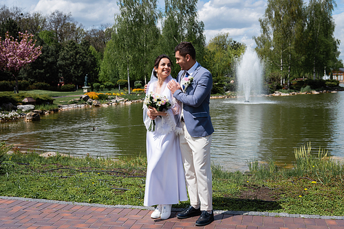 Cheerful bride and groom standing near fountain in park