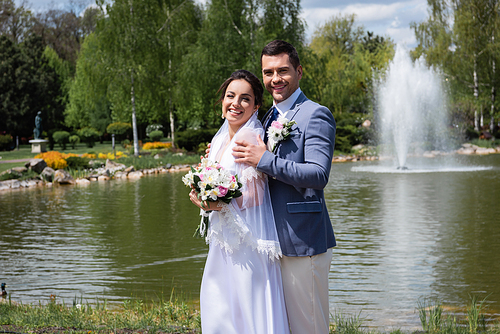 Happy bride and groom standing near blurred fountain in park