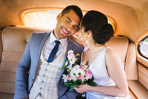 Bride in dress holding bouquet near smiling groom in vintage car