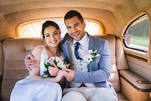 Happy groom embracing bride with bouquet in auto