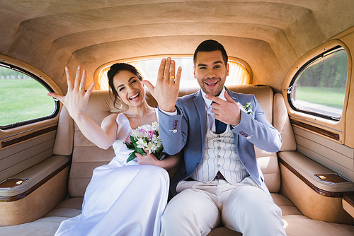Cheerful groom pointing at ring on finger near bride in retro auto