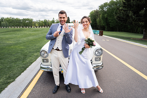 Cheerful newlyweds showing rings near retro auto on road