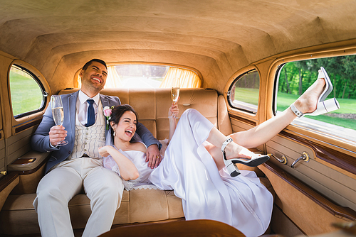 Happy bride holding glass of champagne near groom in retro auto