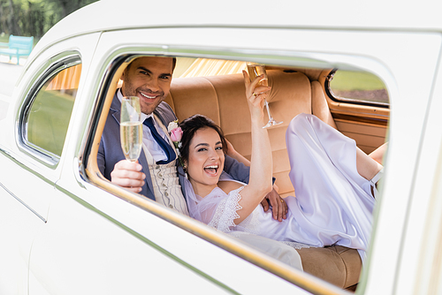 Cheerful groom holding blurred glass of champagne near bride in retro car