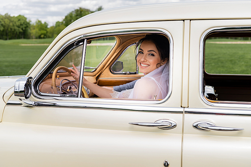 Bride in veil smiling at camera on driver seat of retro car