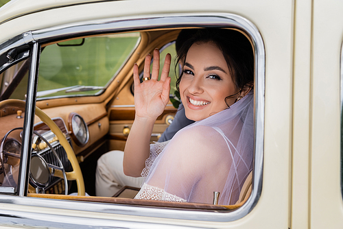 Smiling bride waving hand at camera on driver seat of vintage auto