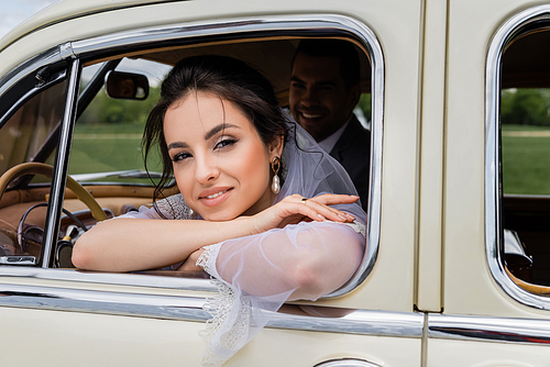 Cheerful bride in white veil  near window of retro car