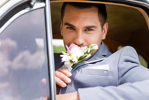 Elegant groom smelling flowers of boutonniere in retro car