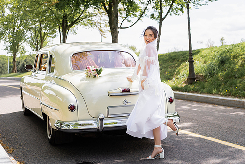 Happy bride standing near bouquet on vintage car on road
