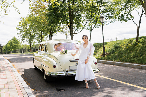 Smiling bride in dress and veil standing near retro car