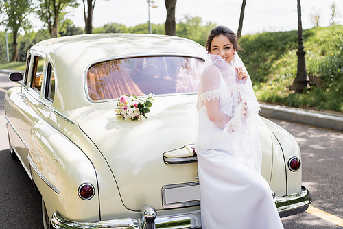 Brunette bride holding veil near vintage car outdoors