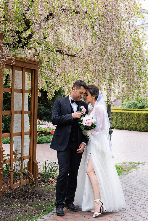 Side view of young newlyweds standing near blooming tree in park