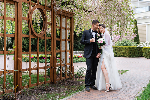 Cheerful groom standing near bride in wedding dress and blooming tree in park