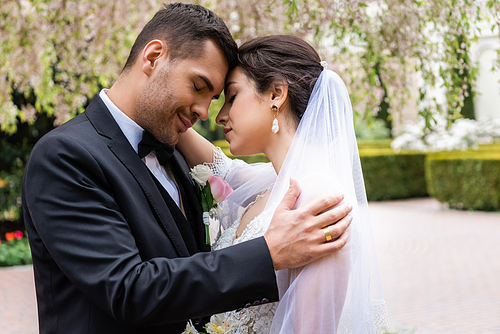 Side view of groom embracing young bride with closed eyes in park